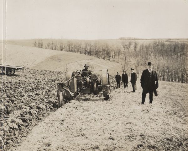 Man driving a La Crosse Happy Farmer tractor plowing in field up a hill with several men in long coats, suits, and hats watching nearby.