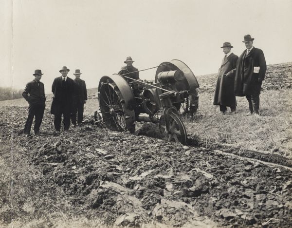 Front three-quarter view of a man driving a La Crosse Happy Farmer tractor in a field plowing. Several men in long coats, suits, and hats are standing on either side of the tractor.