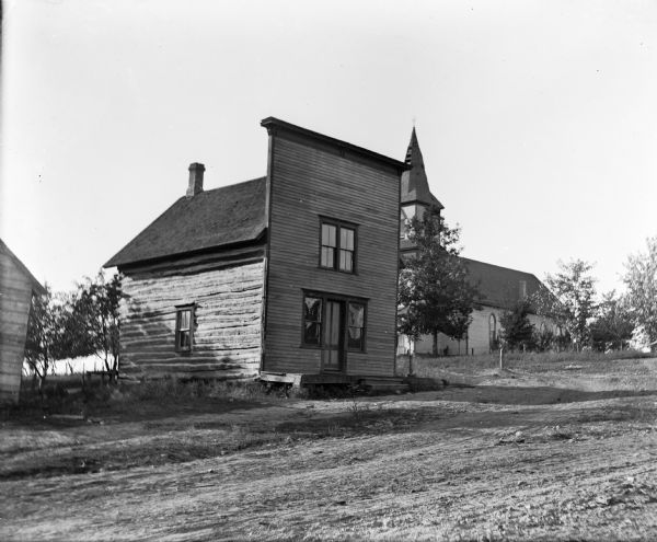 Exterior view of the log building which served as the post office in Loreta (formerly known as Loreto) until January 31, 1907. There is a church in the background.
