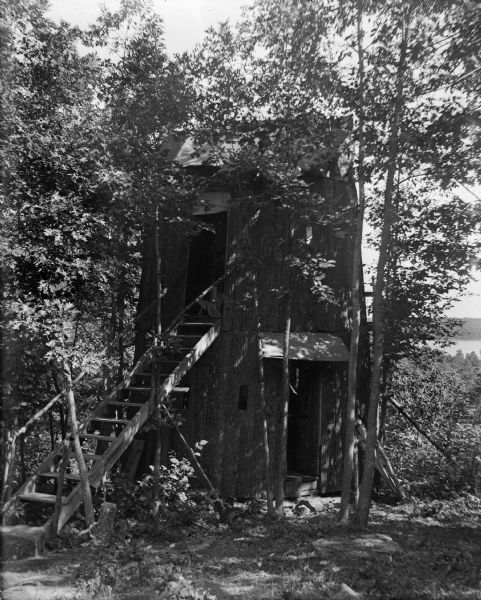 Exterior of bark-clad cabin near Devil's Lake. The cabin has wooden steps leading up to the second floor and is surrounded by trees. The lake can be seen in the background.