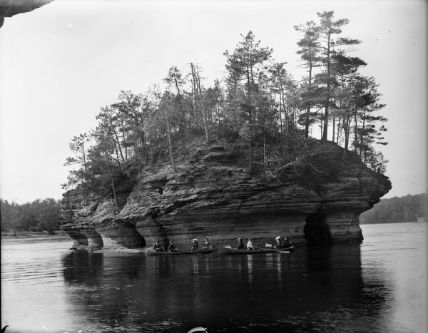 View across Wisconsin River of men and women in canoes at the base of Lone Rock at the Dells.
