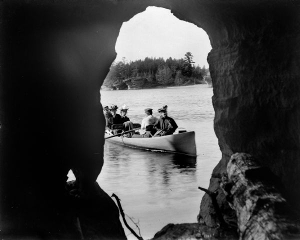A man paddles a canoe with four women passengers, as seen from inside Lone Rock Cave at the Dells. The far shoreline can be seen in the background.