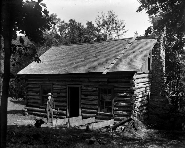W.H. Canfield stands in front of a log cabin. There is a brick chimney on the right side of the building.