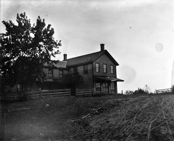 View up dirt road or track towards a two-story L-shaped frame house with a fence along the front. The right end of the house has a roof suspended over the first floor porch or landing, with double doors and two large windows beneath.