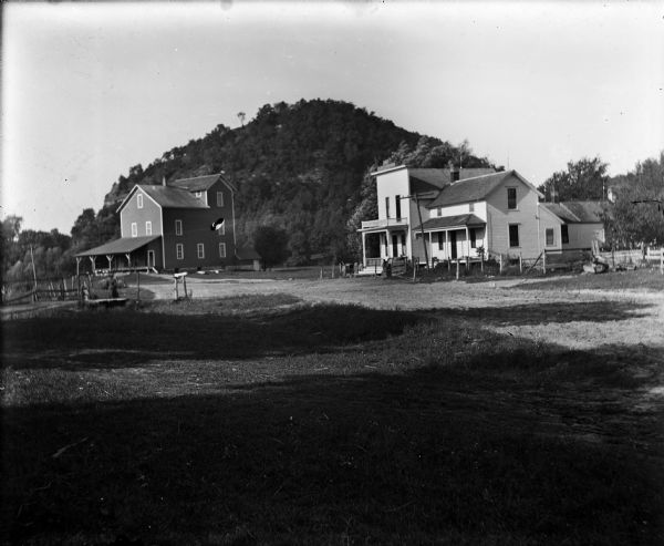 A mill, left, and a commercial building with attached residence stand in front of a wooded bluff. A woman crosses a small bridge near the mill.