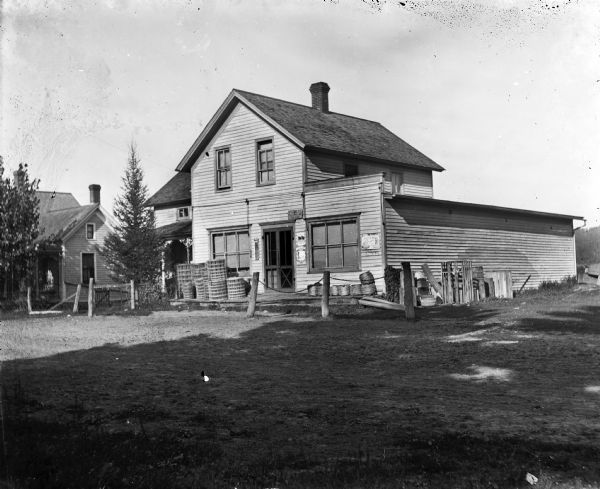 Exterior view of a storefront with rolls of wire and barbed wire fencing materials on the front porch. The store was formerly the post office for Dwyer. A portion of a house can be seen on the left.