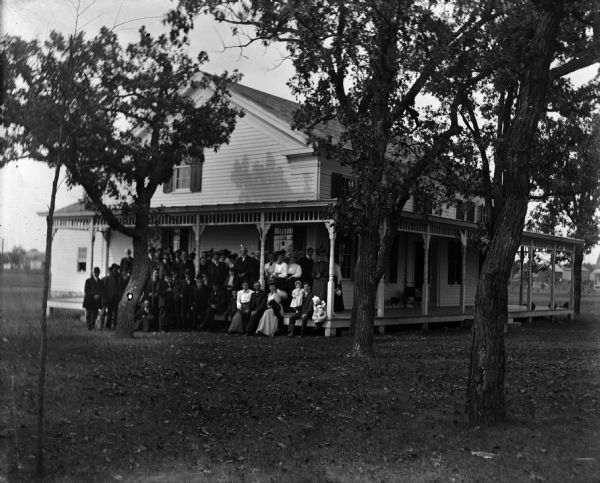 View across yard of house with large group of people gathered on porch posing for photograph.