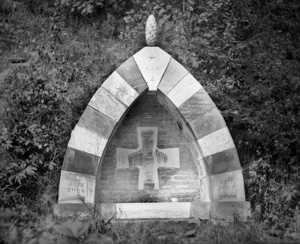 Brick and stone religious fountain in the shape of a Gothic arch, with a cross in the center and a pinnacle on top. The cross is engraved with the words, "Christ said I Thirst / Brother or Sister /  Think of / Him and Drink." A ceramic cup sits on the base of the fountain.