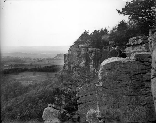 Side view of a woman sitting on a rock ledge on the face of Gibraltar Rock bluff. Hills, fields and the Wisconsin River are visible far below in the background. There are trees growing on the top and face of the bluff.