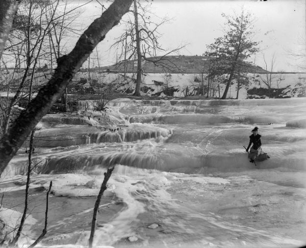 Elevated view of a woman holding a walking stick and wearing a hat and fur coat with a fur stole sitting on a ledge of ice at Skillet Falls. Bare trees frame the scene. There are patches of snow and tree stumps on a hill in the background.