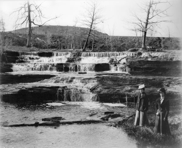 Two well-dressed women stand at the foot of Skillet Falls.  The trees in the background are bare; the hill behind is covered with tree stumps.  The hill is known locally as Mount Baldy.