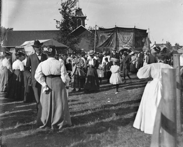 Visitors to the Sauk County Fair walk by sideshow attractions and concession stands while others stand and converse.  The "Grand Electric Palace," a "$10,000 attraction" stands in front of one of the exhibit halls. There is a curved fence on the right.
