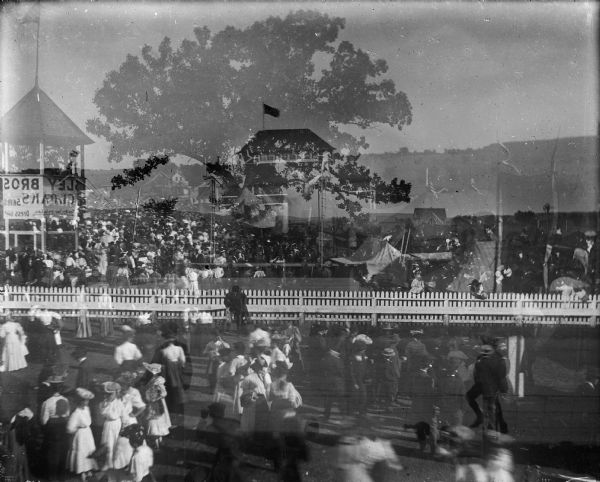 A double exposure of a landscape featuring a very large tree and farm houses superimposed on an image of buildings and crowds at the Sauk County Fairgrounds. A banner advertising a clothier is hanging on an elevated bandstand or observation tower on the left. A picket fence crosses the center of the scene.