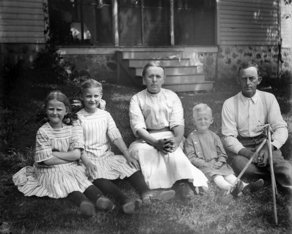 A family group of mother, father, two daughters and son sit on the ground in front of a house with a stone foundation.  A tripod arrangement of a toy shovel, rake, and hoe stands in front of the father.