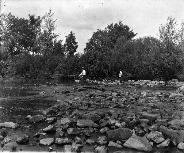 Three men fish with long cane poles at a rock lined stream.