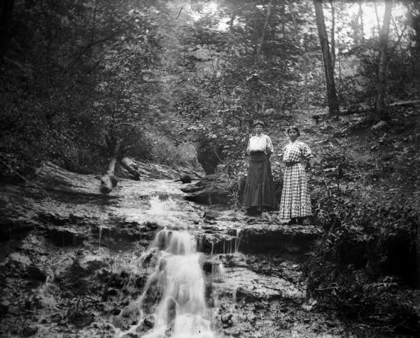 Two women stand beside a waterfall "downstream from the crag."