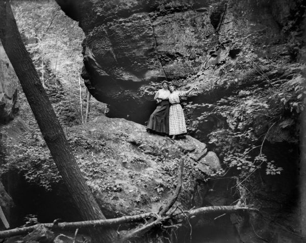 Two women wearing long dresses pose on a large boulder at the base of a rock formation in a shady glen.