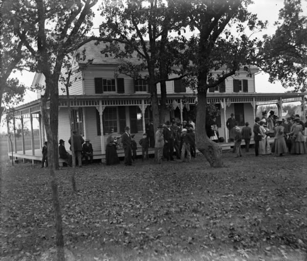 The land and house were donated to the village. The club house is still in use (1964) as a publicaly owned building for recreation and meetings. Image shows a large group of people gathered on the porch and on the yard in front of house. American flags decorate the porch.