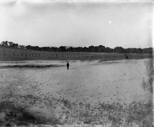 A well-dressed man stands on barren, sandy soil identified as an Indian village site near Okee. Near him the soil is stained darker than the surrounding area. There is a field of corn shocks in the background.