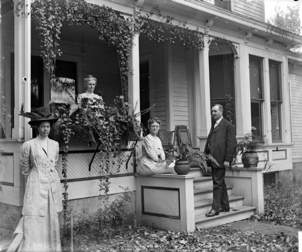 Three women and a man posed on and around the porch of the Monroe House. Potted plants, a flower box, and vines grace the porch.