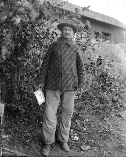 Mr. Kleinlein poses outdoors with a photograph in his hand. There is a large building on a hill in the background.