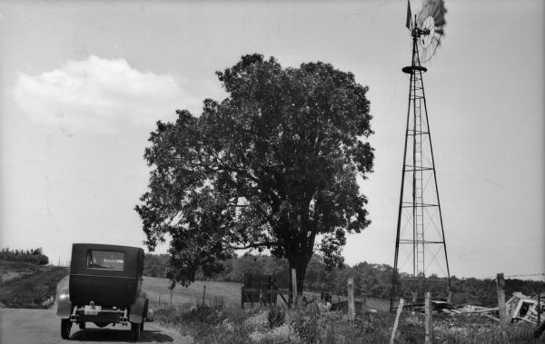 A car and driver stop on a country road near Mt. Horeb at the site of the Holton Tavern.  A windmill and tree stand on the site and there is debris at the base of the windmill.