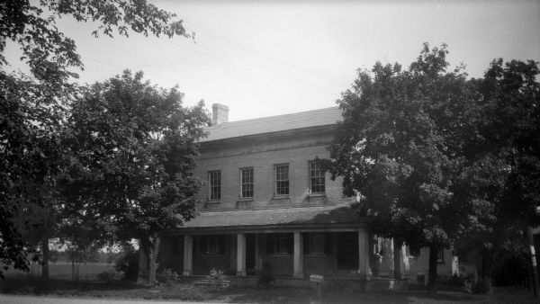 A view of the front of large house with an open front porch, a front yard with mature trees, and a field beyond. The house features six over six window sashes and brick dentil molding.