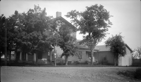 A side view from road of large brick house with an open front porch, surrounded by trees. There is a a rear wing and attached garage or shed.