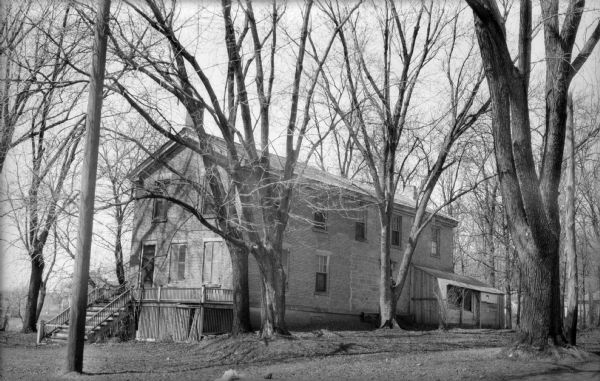 The Plow (Plough) Inn, 3402 Monroe St., surrounded by trees.  The stone section on the right is the original farmhouse. The brick section on the left was added in 1858 by John Whare and his wife Isabella.  He was a glass blower from England.  They named the Plough Inn for the ploughs they sold from the side yard. There are a flight of steps up to the landing / porch and the front door. A small, shed-like structure is attached to the right side of the inn. Other buildings, perhaps houses, can be seen in the background.