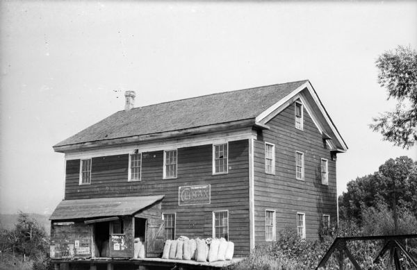 The Loganville Mill, built in 1861. The owner was Joseph Mackey and the carpenter was Billy Palmer. Sacks of feed are sitting on a loading platform. There is a sign for chewing tobacco painted on the building.  A portion of a bridge is on the right.