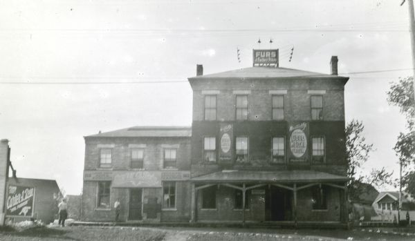 A front view of the Layton House, which was originally an inn. Signs on the building indicate that at the time of the photograph it was the business of Carter A. Reins, furrier.  Two men stand in front of the building, and a person or persons lean from a second floor window.