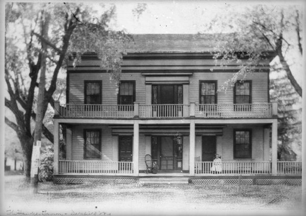 Front view of Hawks Tavern, also known as Hawks Inn, with a person sitting on the porch. There is also a second floor porch. Posters are posted on a pole in front of the building.