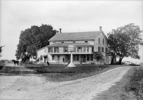 View from fork in road of Martin's Tavern in Chamberlain. There is a horse and carriage with driver on the left. Three men sit or stand in front of the tavern. There is a multiple chambered bird house on a post in the yard.