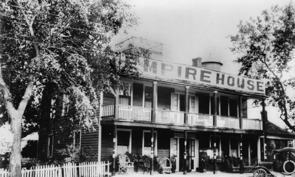 A view of the front of the Empire House Hotel, showing the porch and balcony. There are many wicker chairs on the porch, and there is an automobile parked on the right in front of the hotel. There appears to be a widow's walk and a small, conical-roofed building on the roof.