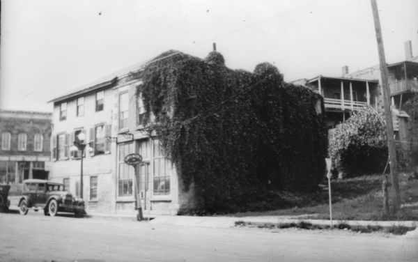 View across street of several commercial buildings in a downtown area. The stone building in the foreground is nearly covered by vines and is identified as the City Hotel. An automobile is parked in front.