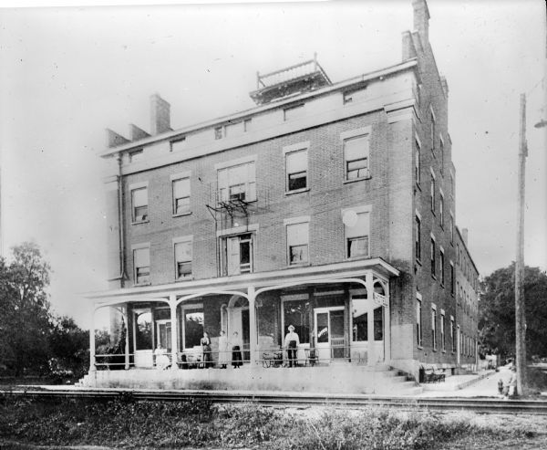 Denniston House, P.J. Schnorrenberg, proprietor. View across railroad tracks of the hotel entrance with porch. A man and three women are standing on the porch. A widow's walk is on the flat roof of the building.