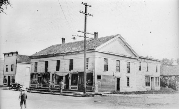 View across street of a young boy in overalls crossing the street in front of a large frame commercial building with windows and awnings, possibly a hotel.  There is a man walking off the curb in front near a large telephone pole and suspended streetlight. An automobile is parked on the street on the left near another wood frame commercial building.