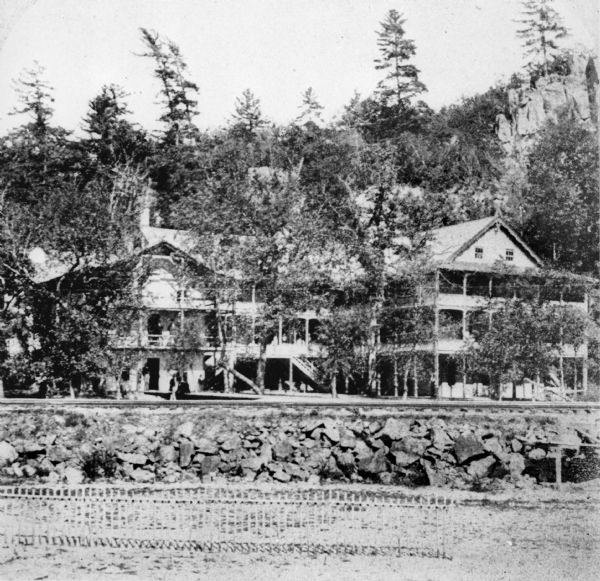 A copy of an original photograph by H.H. Bennett of the Cliff House Inn at Devil's Lake as seen from the beach. Abraham Lincoln is reported to have slept here. There are large porches on the front of the building.  A bluff is in the background and railroad tracks run between the inn and the beach.