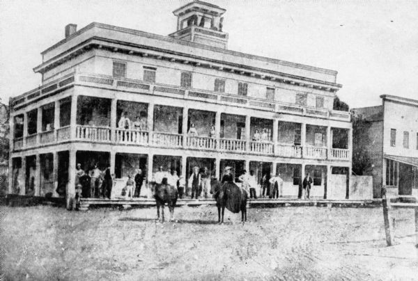 A woman on horseback poses with another horse in front of the three-story Western Inn as people look on from the porches.  There is a large belvedere on the roof.