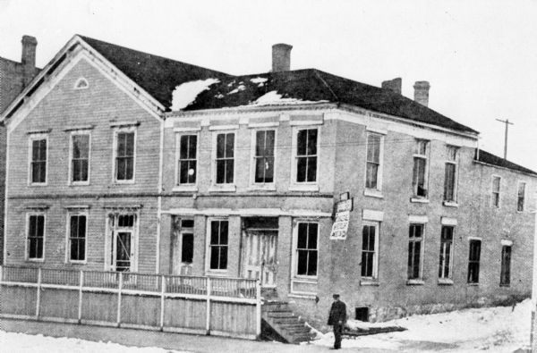 A man stands near the wooden steps of the Wisconsin House, a wood frame building with a large brick wing. There is snow on the ground and on the roof.