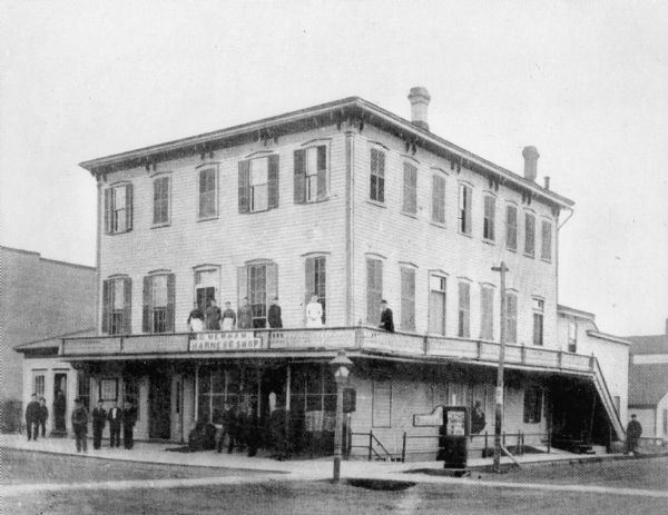 Men stand on the street level porch and women on the second floor wrap-around balcony of the Alba House. A sign on the railing advertises a harness shop.