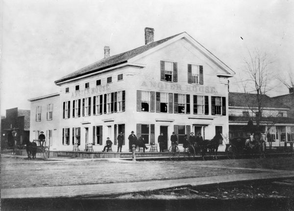 Men with horses and buggies pose in front of a three-story wooden building identified as the Angier House by a sign painted on the clapboards. Women look out the upper story windows.