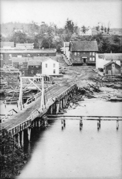 Elevated view of a small town with a river and bridge in the foreground. High water has washed away the road and river bank at the far end of the bridge. There is a church on the hill in the background.