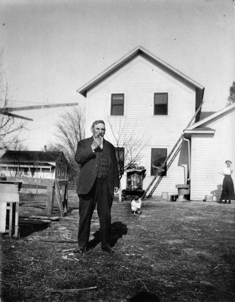 Herman Rafoth, smoking a pipe, stands behind a two-story wooden house.  Rafoth was the driver of a stage held up by Raymond Holzse on May 8, 1880.  A woman in the background shields her eyes from the sun. There is a chicken walking in the yard.