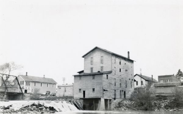 The dam and four-story wooden mill on the Baraboo River.  A bridge crosses the river on the left. There are houses and commercial buildings in the background.