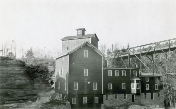 View across lake of a four-story wooden mill with two wings, one with an oriel window. There is a building with a cupola, probably a granary, behind the mill. Large rock formations are on the left, and a high trestle bridge is on the right.