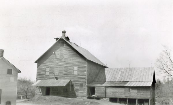 A large wooden building with side wing and covered porch, this mill was built in 1853-4 by Newell Dustin. There is a cupola behind the chimney. According to the photographer's notes, Dustin "borrowed (a) team from William Wilcox to haul millstones from Milwaukee."