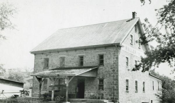 Exterior of stone mill. An automobile of the period is parked under the front portico, and there is a wooden wing in the rear. A sign in front advertises "Middlings for Sale," a sign on the side of the building says, "Roller Mill."  This mill was built about 1858 by Oscar Mohr and Dr. Francos Fisher.
