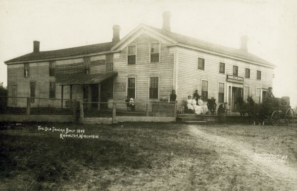A photographic postcard of "The Old Tavern" built at Knowlton in 1849.  It is a large wooden classical revival style building. A dog joins a group of people sitting and standing on the porch. A man sits on bales of hay on a horse-drawn wagon in front of the building on the right. A large sign over the door reads "Knowlton House." The side yard is fenced with chicken wire.