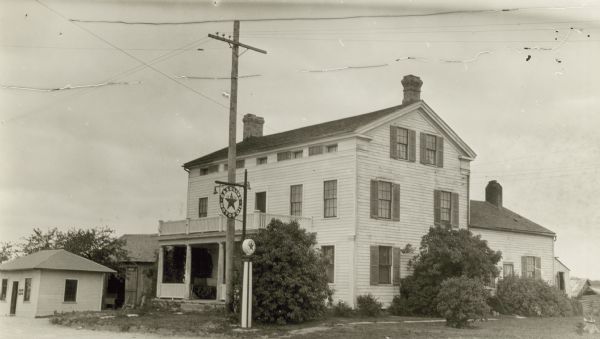 A large wooden classical revival style building with rear extension identified as the Dousman House, or tavern. "[It] stands a few miles east of Waukesha, where the road branches into Grand Avenue, Milwaukee." A gas pump and a sign for Texaco gasoline and motor oil are in front of the building.